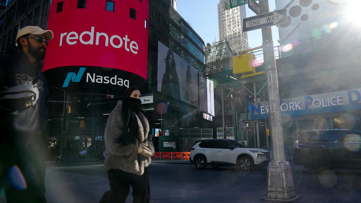 Two pedestrians walking in Times Square, with a large digital billboard displaying 'rednote' and the Nasdaq logo in the background, alongside street signs indicating 'W 43rd St' and 'ONE WAY', and a New York Police Department sign visible nearby.