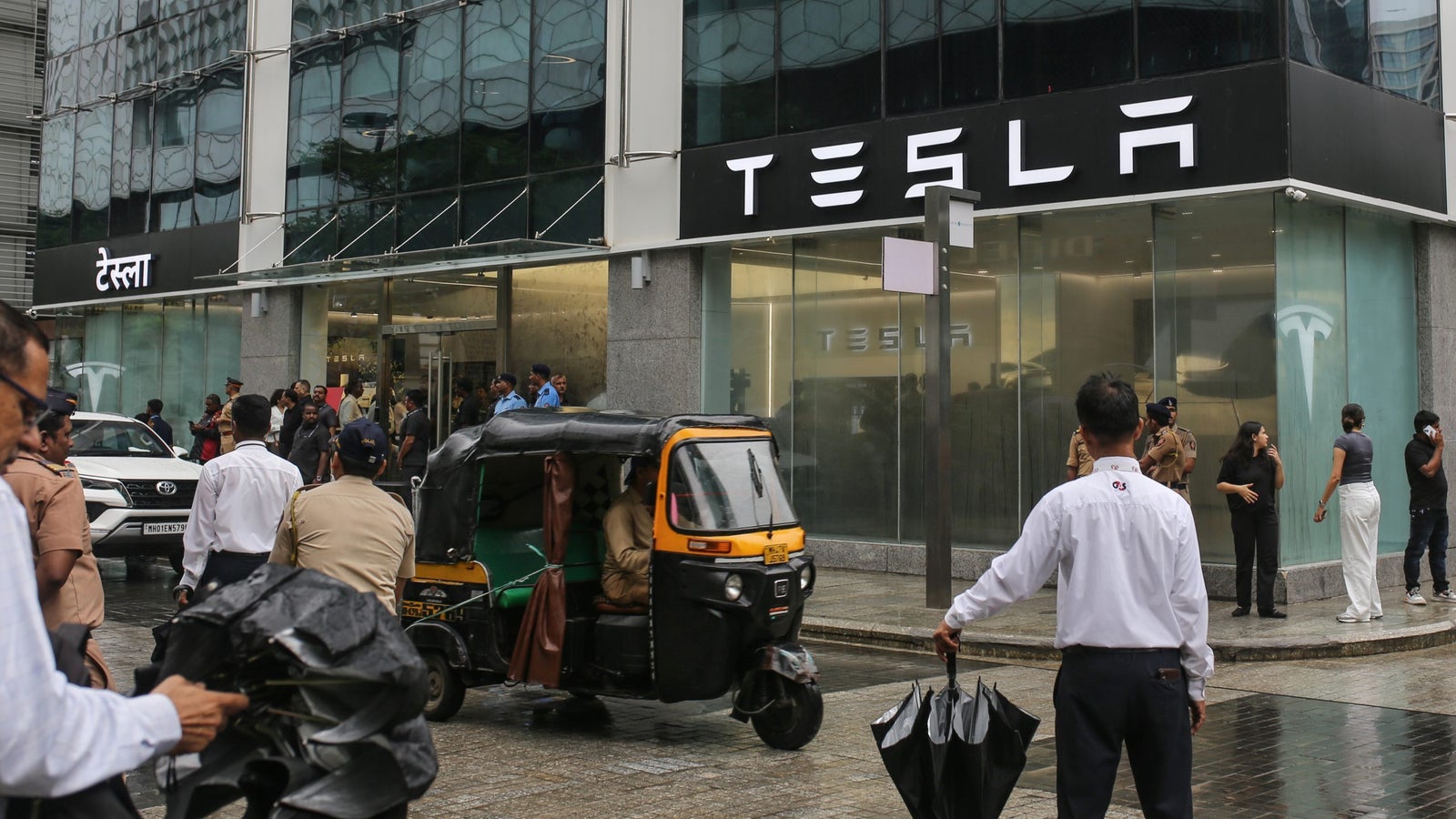 A busy street scene outside a Tesla store with people, including police officers, gathered at the entrance, while an auto rickshaw drives by under a rainy sky.