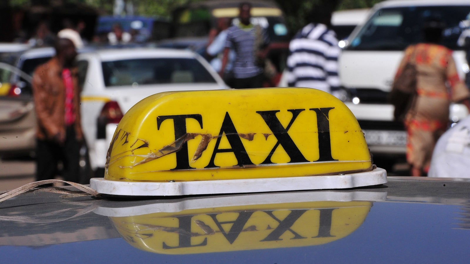 A close-up of a yellow taxi sign on top of a car, with the word "TAXI" prominently displayed in black letters. Blurred background shows people and vehicles in a busy street in Kenya, Nairobi.