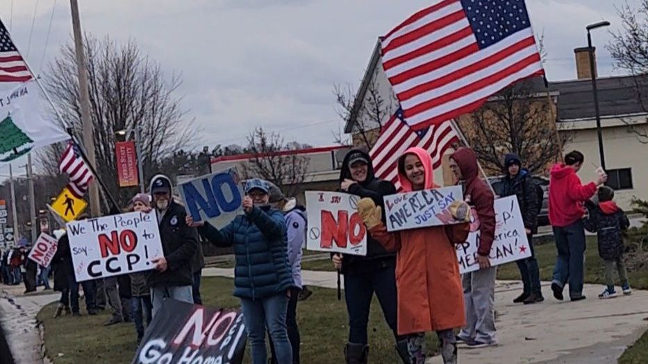 A group of protesters holding signs and American flags during a demonstration, with messages like 'NO CCP', 'We the People say NO', and 'Go Home'. The crowd is dressed in winter clothing, and some people are visibly smiling and posing for the camera.