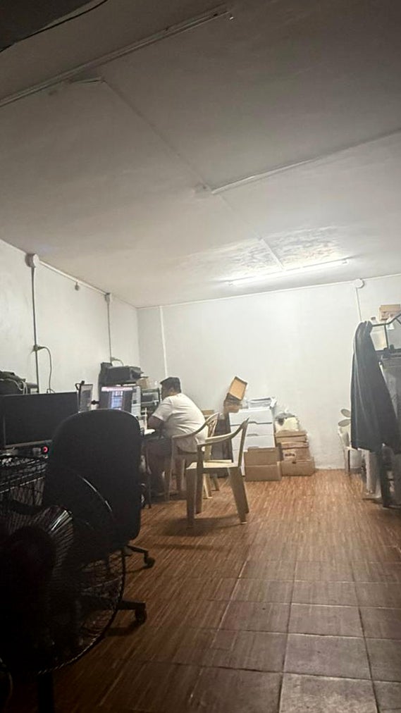A dimly lit room with wooden flooring, featuring a person seated at a desk with a computer, surrounded by stacks of boxes and various items on a table. A standing fan is visible in the foreground.