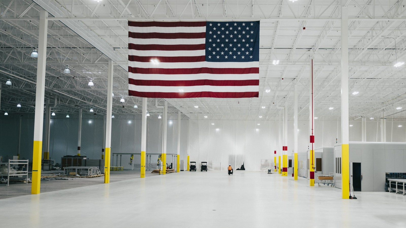 An expansive, empty warehouse interior featuring a large American flag hanging from the ceiling, yellow painted columns, and various industrial equipment scattered throughout the space. The bright lighting illuminates the clean, white floor.