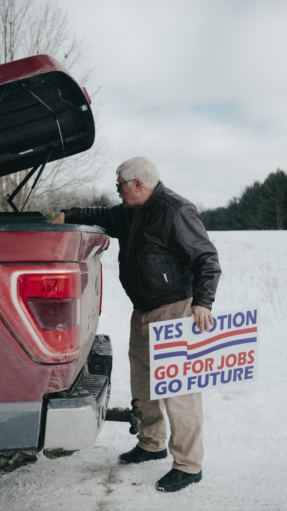 An elderly man with glasses and white hair stands beside a red truck, holding a sign that reads "YES CAUTION GO FOR JOBS GO FUTURE" while looking into the truck bed, with snow-covered ground and trees in the background.