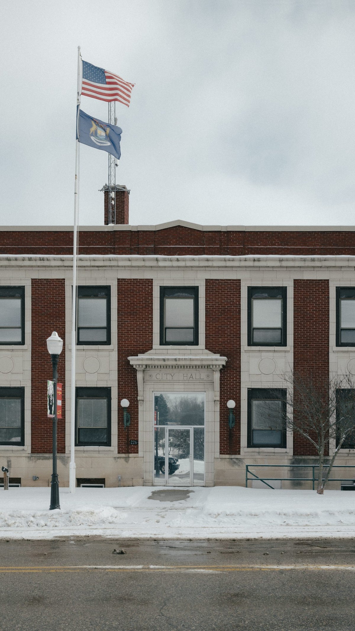 Facade of a brick building labeled 'CITY HALL,' featuring an American flag and a state flag on poles, with snow covering the ground and a clear sky overhead.
