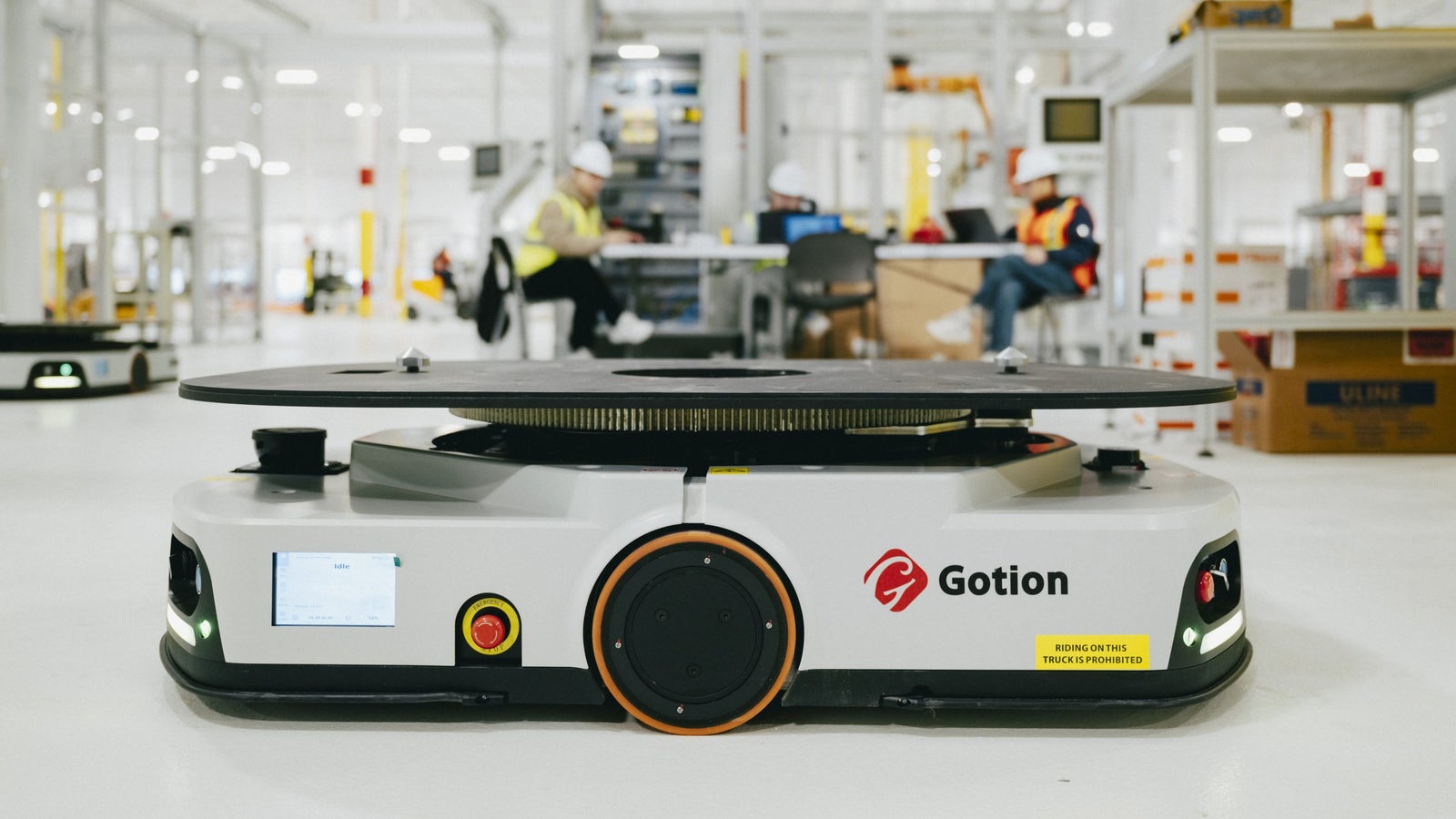 A Gotion-branded autonomous robot with a flat top, positioned in a warehouse environment with workers in safety gear in the background. The robot has a display screen and notable safety features, including a warning label against riding on it.