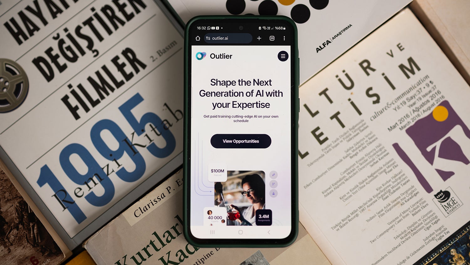 A smartphone displaying a webpage for "Outlier" with text about shaping the next generation of AI, placed on a stack of Turkish magazines and books featuring various titles and dates.