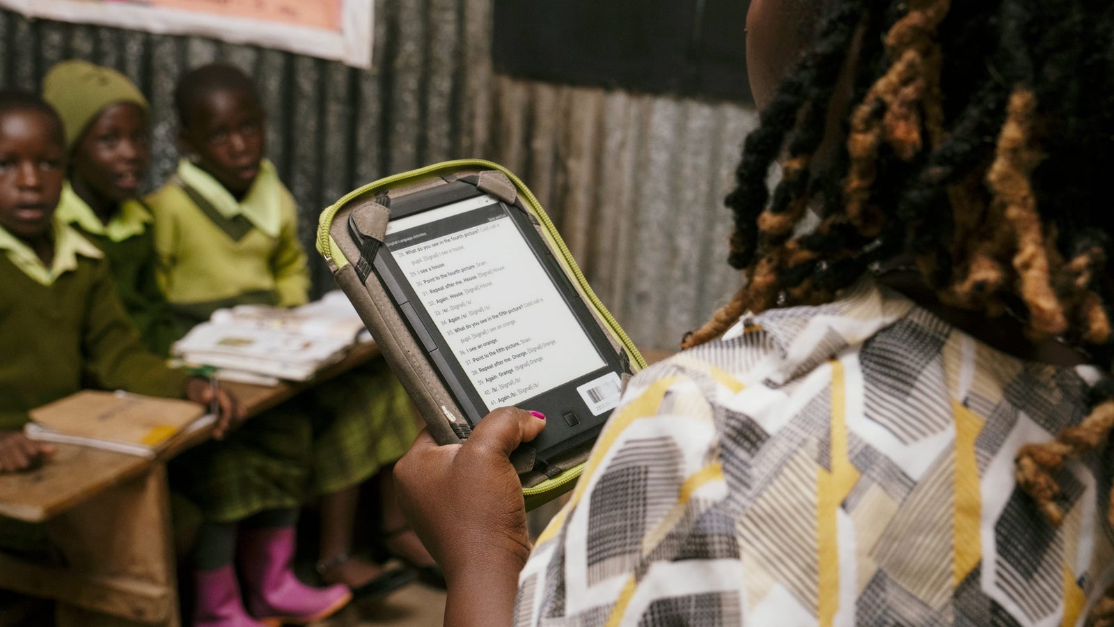A person holds a tablet displaying text, while seated schoolchildren in green uniforms are visible in the background, engaged in their studies.