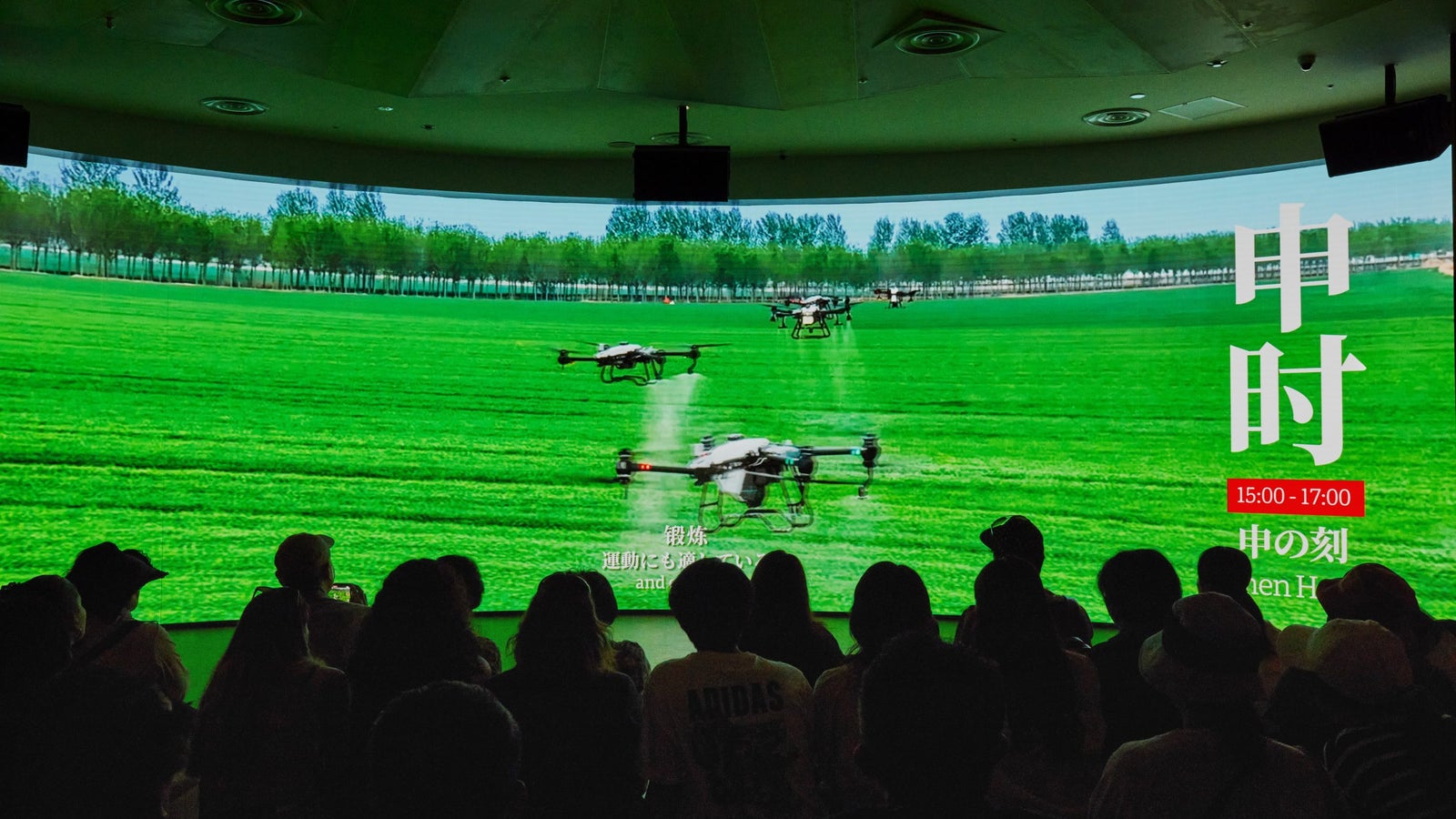 A group of people watching a large screen displaying multiple drones flying over lush green grass and trees in the background, with text in Japanese indicating the event time and title.
