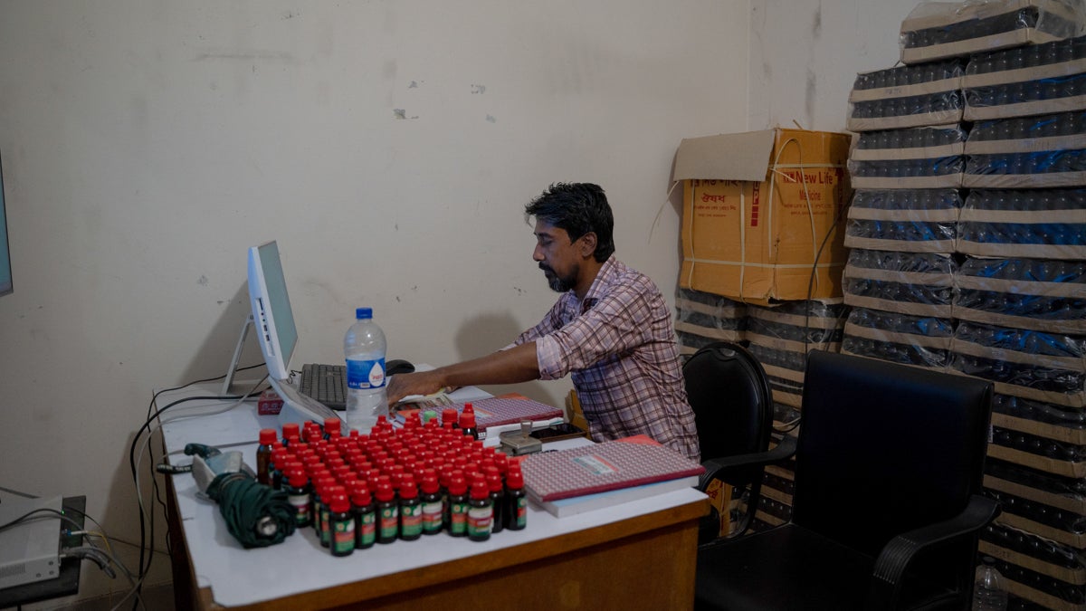 A person works at a desk with a computer, surrounded by bottles with red caps and folders, in a sparsely furnished office space with stacked cardboard boxes in the background.