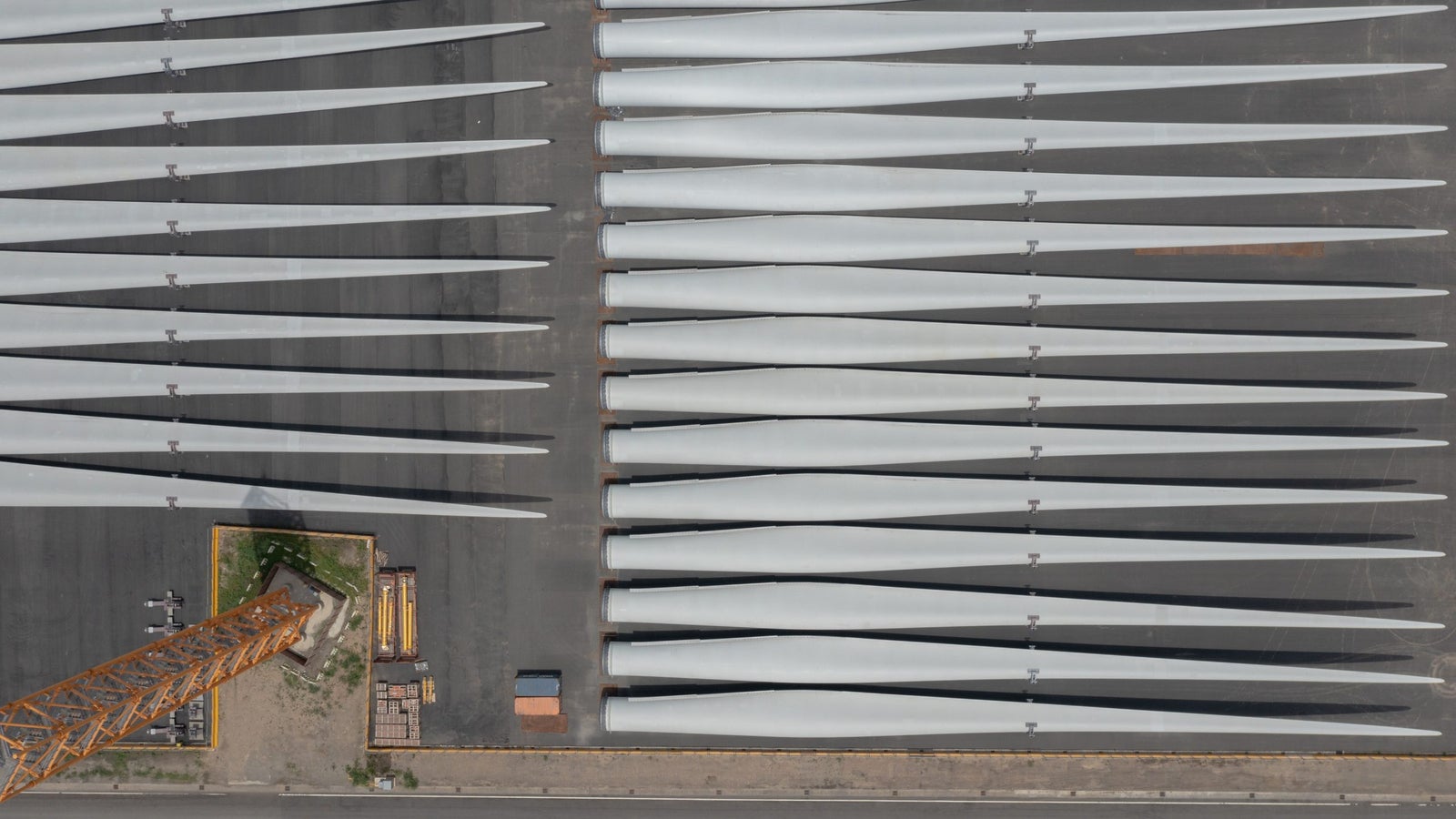An aerial view of a storage area with several large wind turbine blades arranged in parallel rows. In the foreground, a tall orange crane is positioned next to the blades, with a road and a red vehicle visible below.