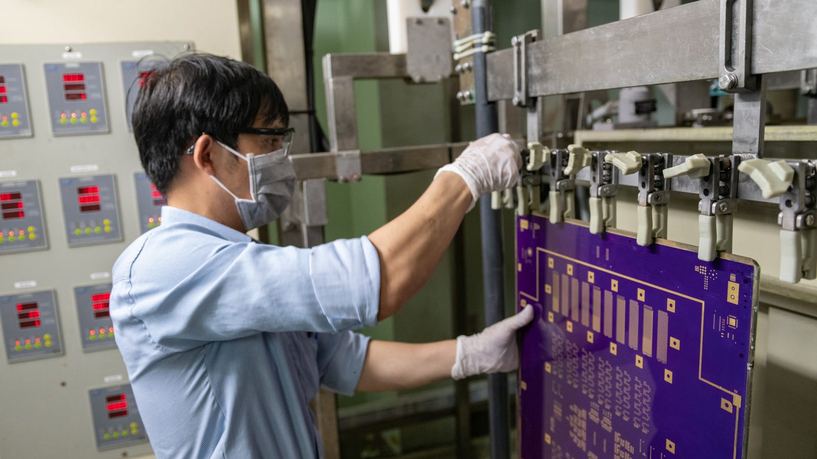 A worker in a blue shirt and mask operates a machine in a manufacturing setting, handling a purple circuit board while wearing white gloves; control panels with red indicators are visible in the background.