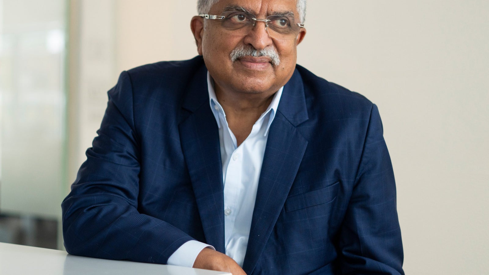 Nandan Nilekani, CEO of Infosys, with gray hair and glasses is seated at a table, wearing a blue blazer over a white shirt, looking thoughtfully to the side in a well-lit, modern environment.