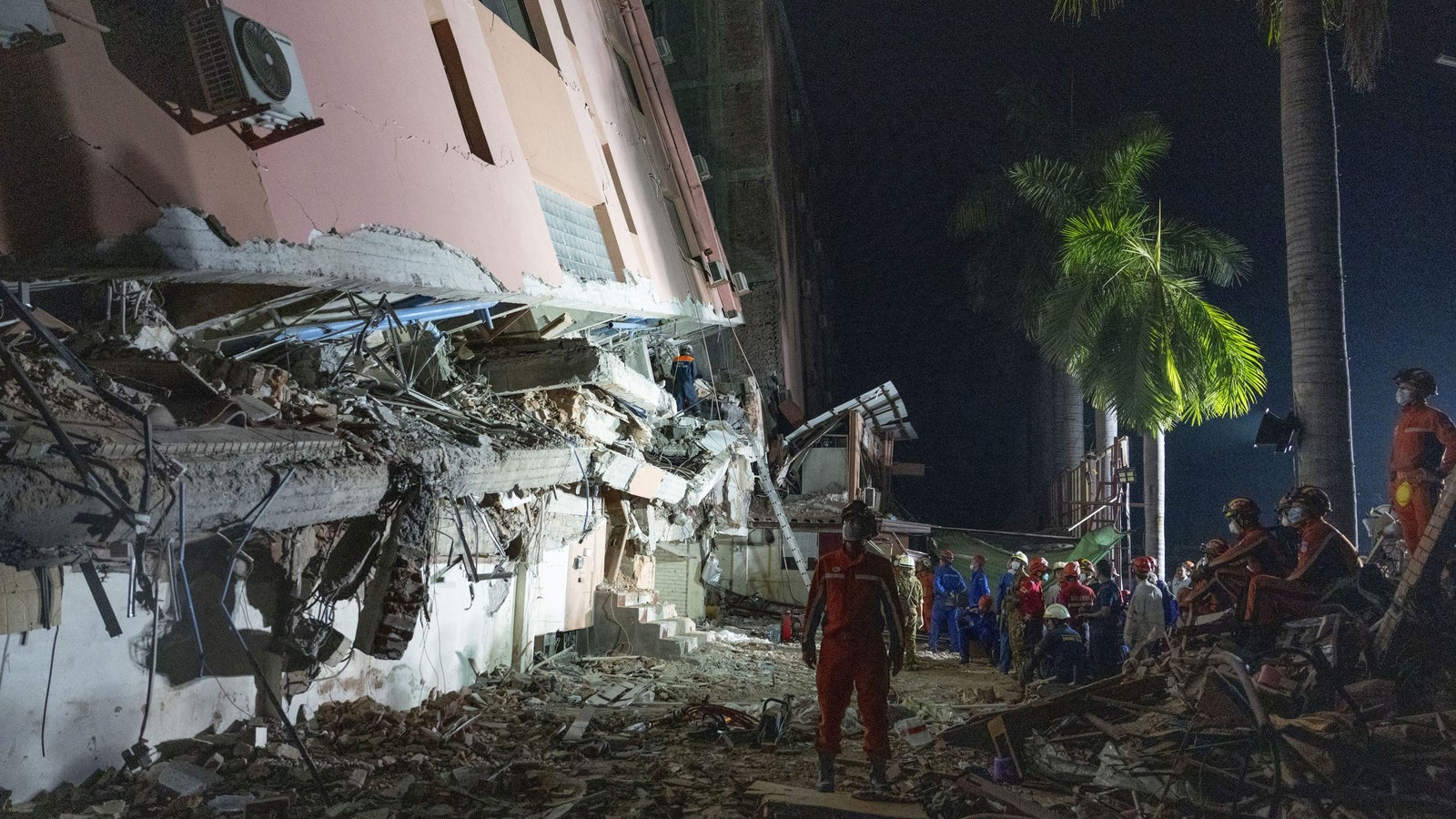 Rescue workers search among the debris of a collapsed building at night, with some wearing helmets and uniforms, while palm trees are visible in the background.