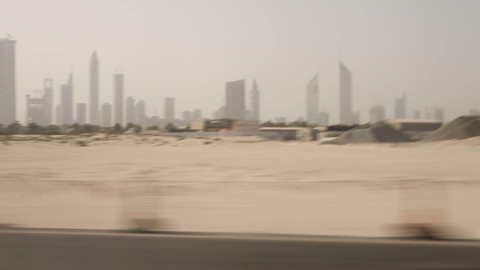 A blurred view of a city skyline featuring tall buildings in the background, with desert terrain and construction materials in the foreground, suggesting movement.