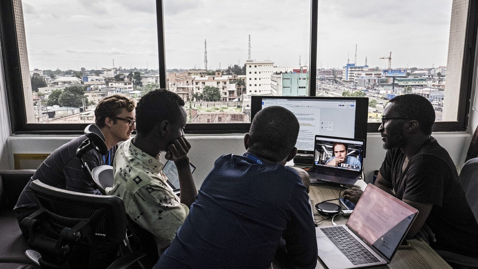 A group of four people in an office setting are engaged in a discussion, with two of them facing a large monitor displaying a video call. The view outside the window shows a cityscape with buildings and telecommunications towers under a cloudy sky.