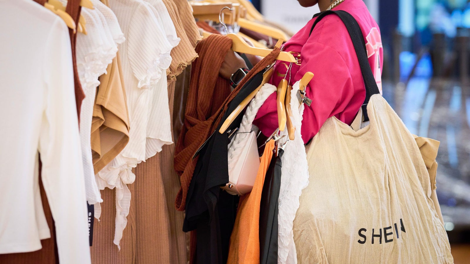 A shopper browsing through a clothing rack filled with various tops in neutral and pastel colors, while holding a large beige tote bag with the brand name 'SHEIN' visible.