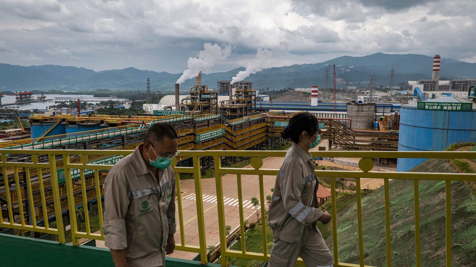 Two workers in beige uniforms and face masks walk along a balcony overlooking a large industrial facility with smokestacks and machinery, set against a backdrop of mountains and a cloudy sky.