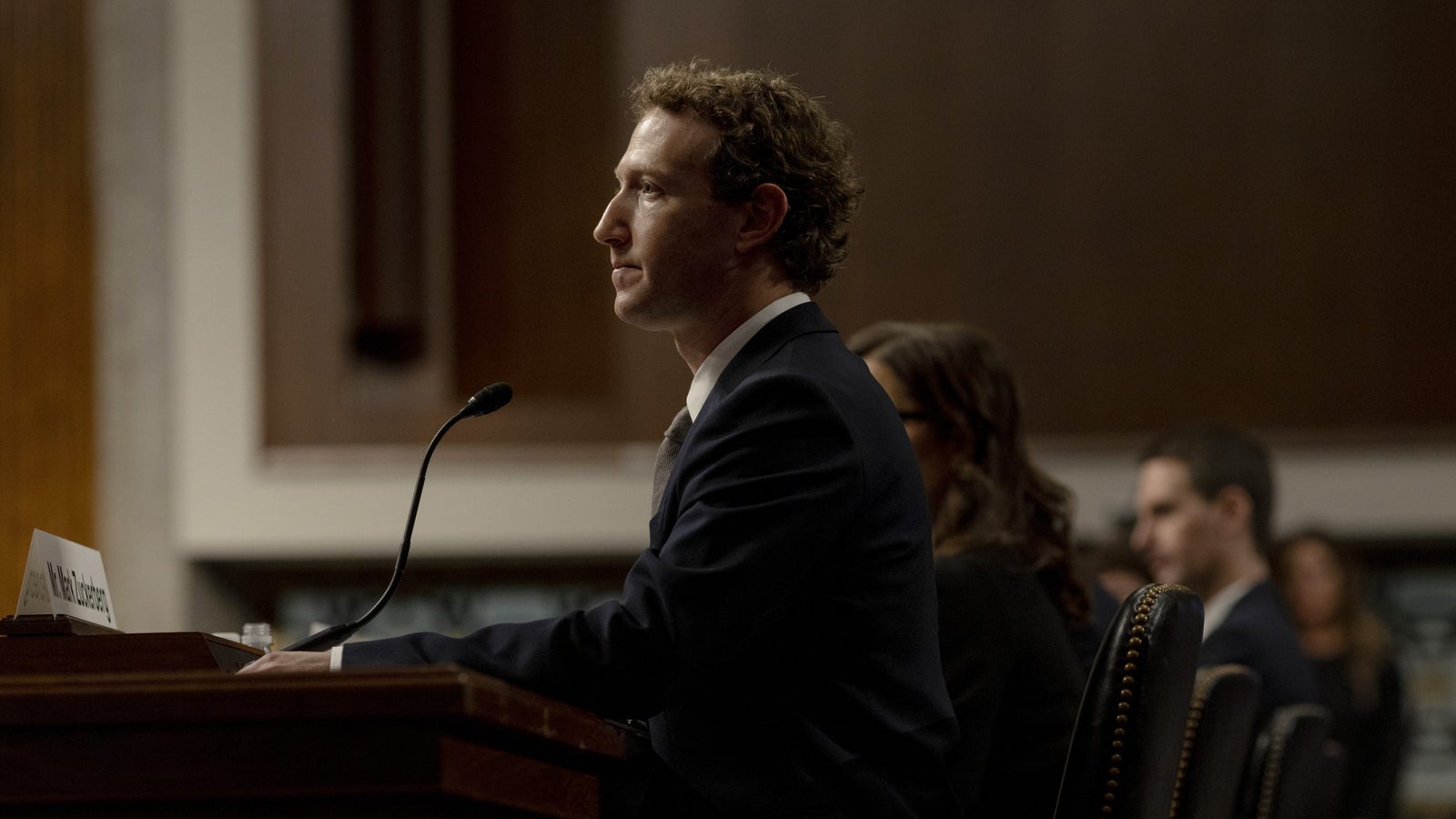 Mark Zuckerberg, chief executive of Meta, in a suit is seated at a podium with a microphone, during a formal event, appearing to be testifying or speaking. There are blurred figures seated behind him in a hearing room. A nameplate in front of him is visible.