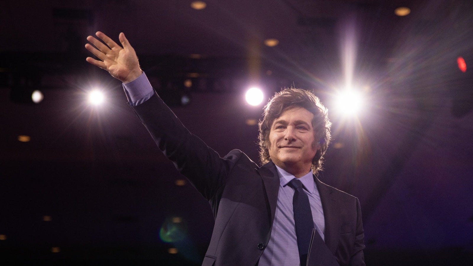 Argentinian President Javier Milei in a suit waves to an audience, smiling confidently, with stage lights creating a dramatic effect in the background.