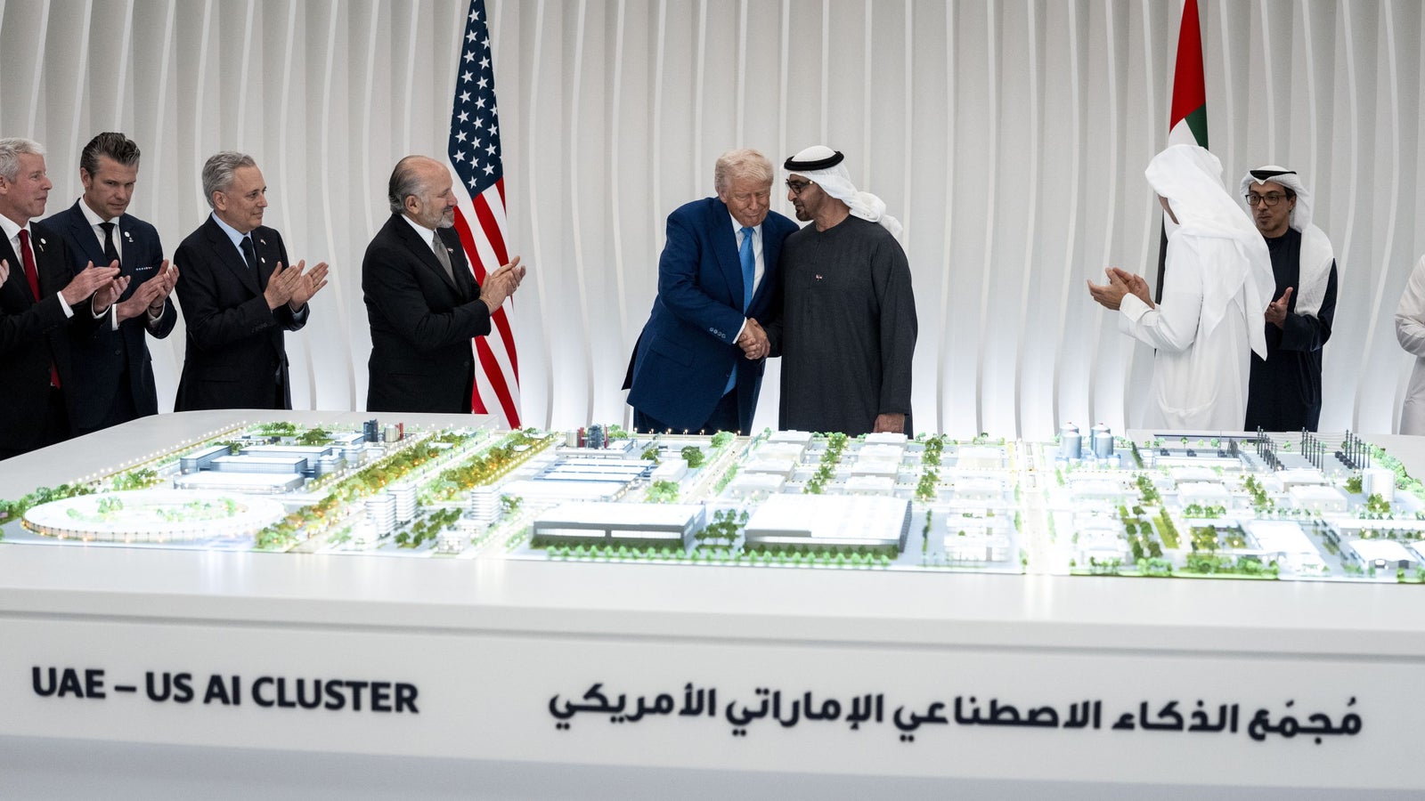A group of individuals, including two men shaking hands, stand around a scale model of a development labeled 'UAE - US AI Cluster,' with a backdrop featuring American and UAE flags. The atmosphere is celebratory, with several people clapping.
