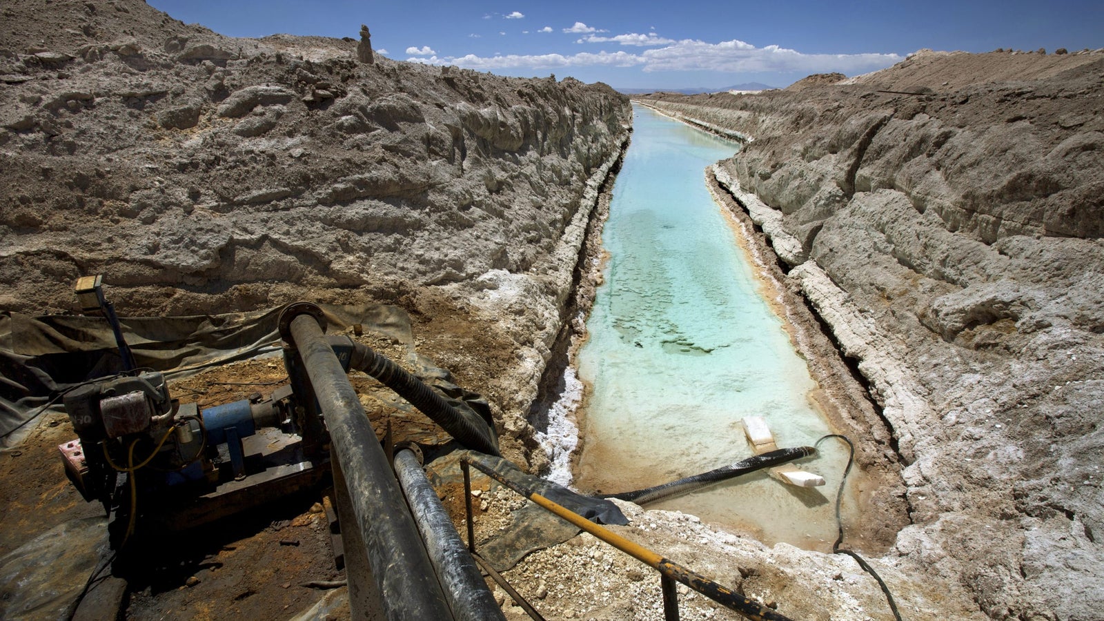 A landscape featuring a narrow, watery channel surrounded by rocky terrain and mounds of mineral deposits, with a mechanical pump in the foreground and clear blue skies above.