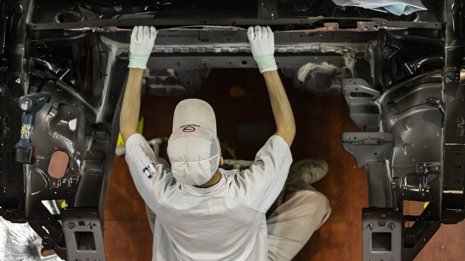 A worker in a light-colored uniform and cap is seated and using both hands to manipulate a metal frame inside a vehicle assembly area, with tools visible nearby.