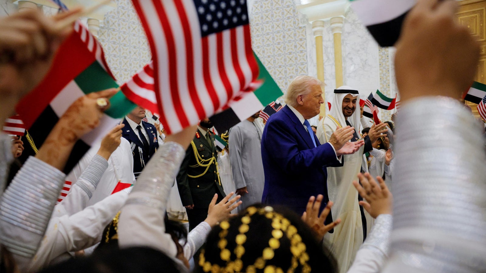 A crowd waves American and Emirati flags while a man in a blue suit interacts with another man in traditional Emirati dress, celebrating in an ornate interior.