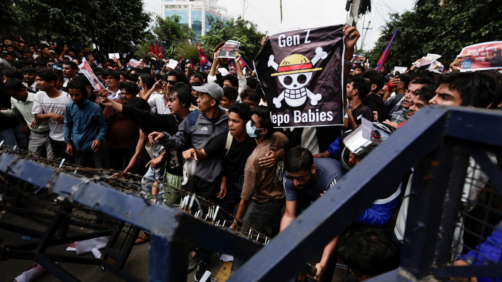 A large group of young protesters gathered behind a barrier, holding up signs; one prominently features a cartoon pirate character and the text 'Gen Z Lepo Babies'. The crowd appears energetic and engaged in a demonstration.