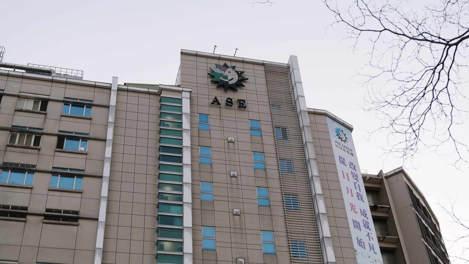 A modern building with blue-tinted windows, featuring the letters 'ASE' prominently on the façade and a large decorative emblem above. A vertical banner with text in Chinese is displayed on the side of the building. The sky above is clear with a faint gradient.