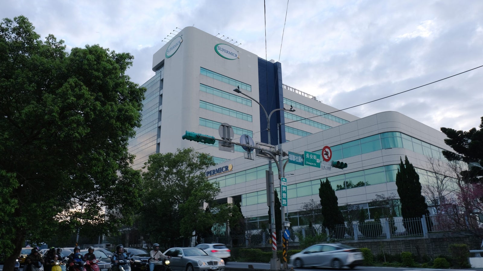 An urban scene featuring a large, modern building with glass windows labeled 'SUPERMICR,' surrounded by greenery and a busy street with motorcycles and cars. Traffic lights and street signs are visible in the foreground against a cloudy sky.