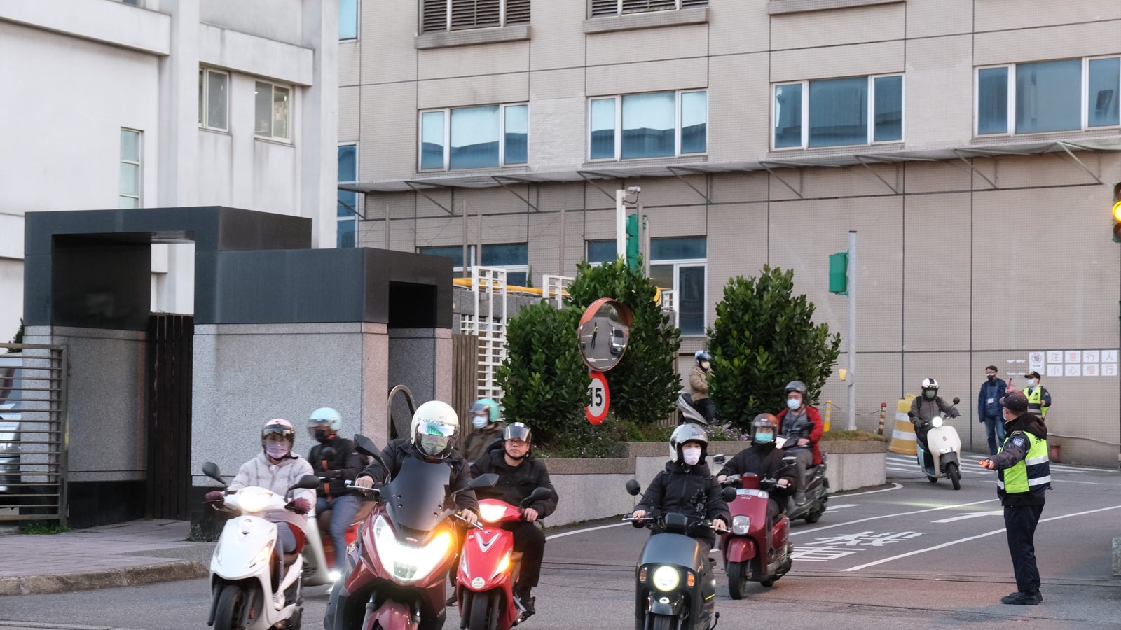 A group of four scooters and motorcycles is waiting at an intersection, with a police officer directing traffic. The scene shows a mix of riders wearing helmets and masks, with a building and greenery in the background. A traffic light indicates stop, and there are road signs nearby.