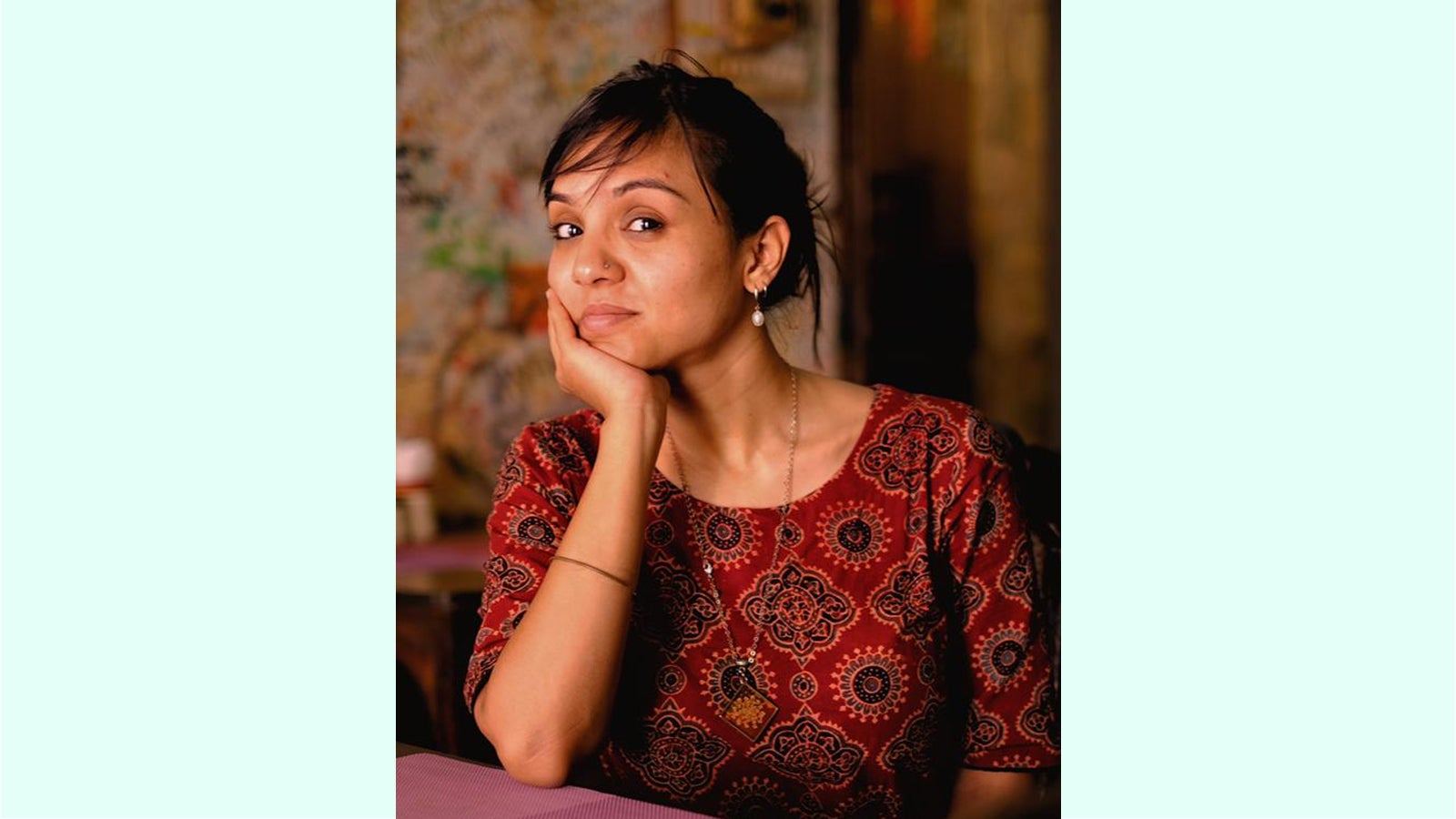 A portrait of Nikita Saxena, contributing editor at Rest of World, with short dark hair and a nose ring resting her chin on her hand, smiling slightly while wearing a red patterned shirt. The background is warmly lit with a blurred decorative setting.