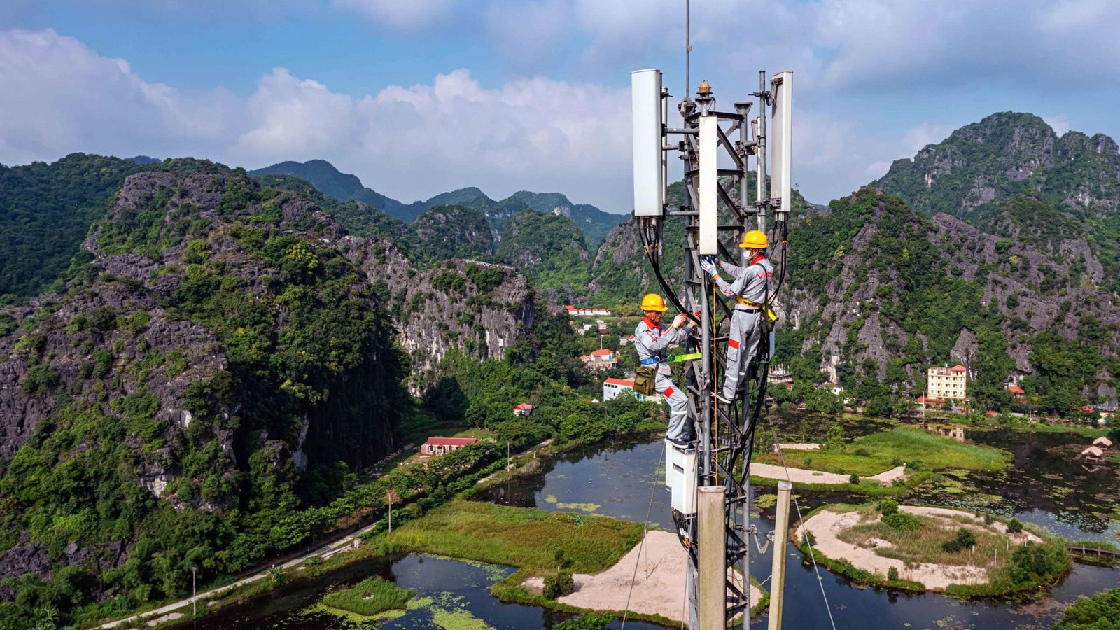Two workers in safety gear and helmets are climbing a telecommunications tower amidst a lush green landscape with residential buildings and a canal visible below.