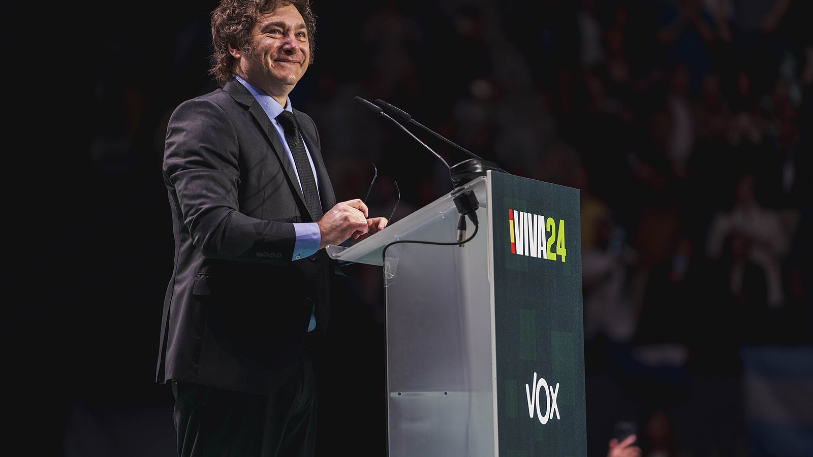 A man with curly hair, wearing a suit and smiling, stands at a podium with a sign that reads 'VIVA24' and 'VOX' during an event, with a blurred audience in the background.