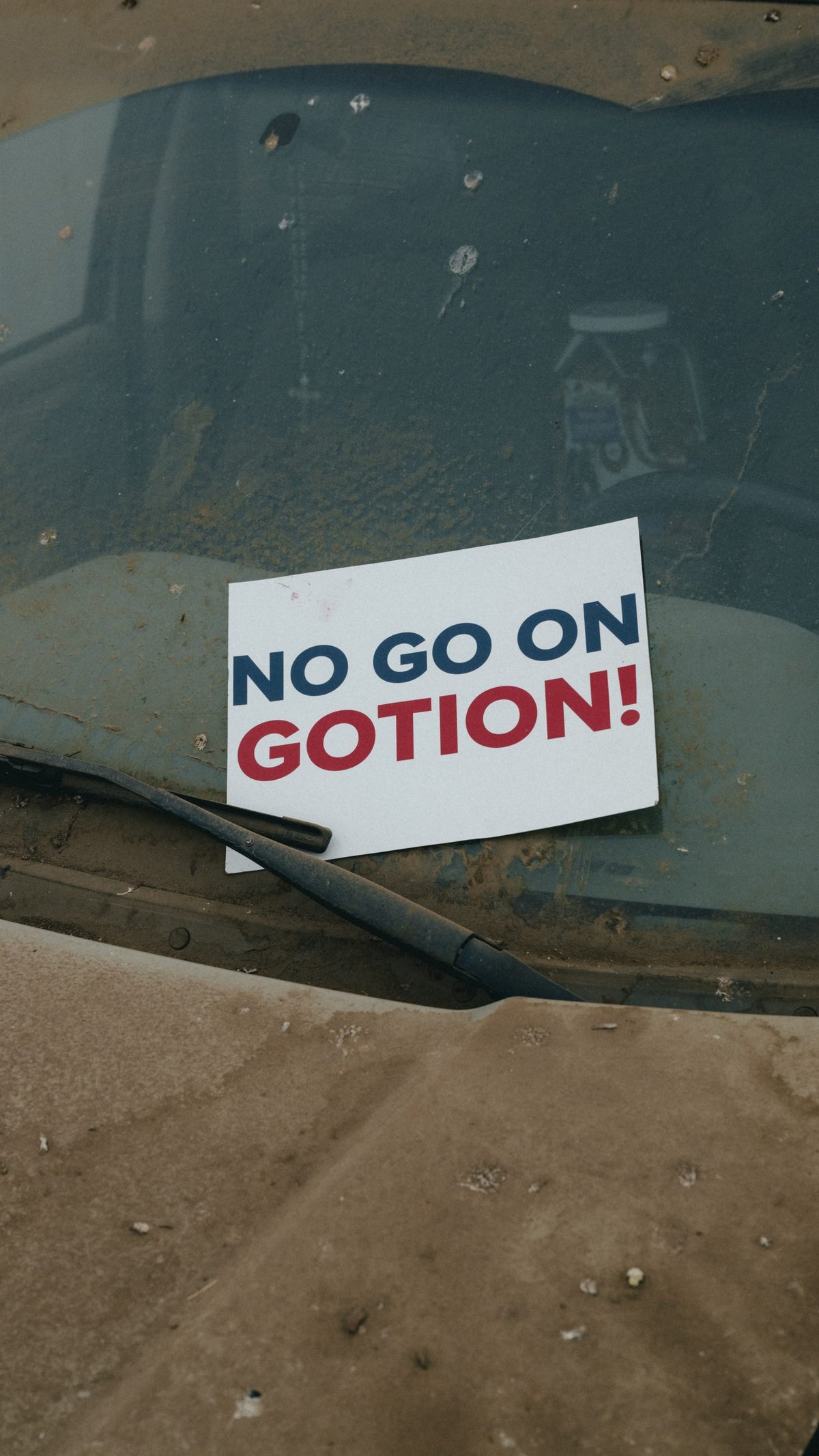 A weathered car windshield with a dirty surface, showing a sign that reads 'NO GO ON GOTION!' in bold red and blue text.