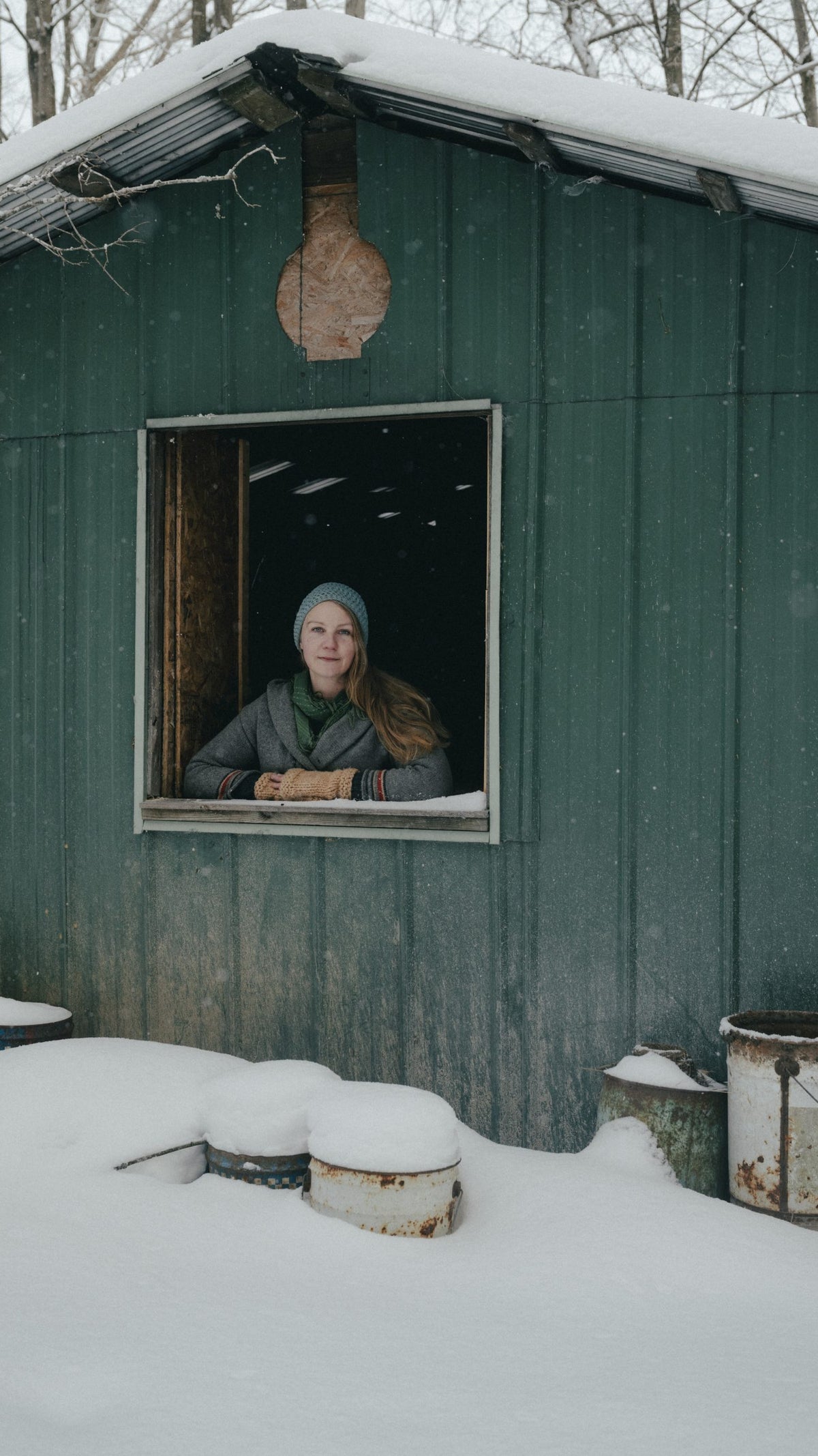 A woman with long hair and a knit beanie leans out of a window of a green metal building covered in snow, with snow piled around various weathered barrels in the foreground.