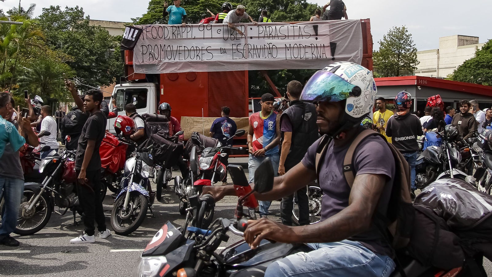 A group of protestors gathered around motorcycles, holding a large banner criticizing delivery companies like Rappi and Uber. Some individuals are on the trucks while others appear to be actively engaged in discussions. The scene is set in a public space with trees and buildings visible in the background.