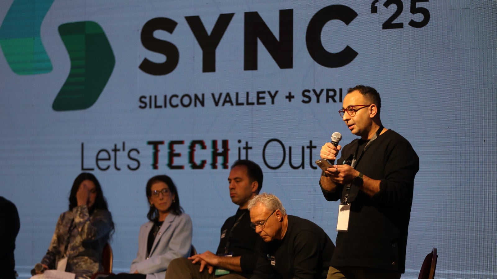 A man in a black sweater holds a microphone while speaking on stage at a conference, with a large screen behind him displaying the logo for "SYNC '25 Silicon Valley + Syria" and the phrase "Let's TECH it Out." Other participants sit on stage, engaged in the discussion.
