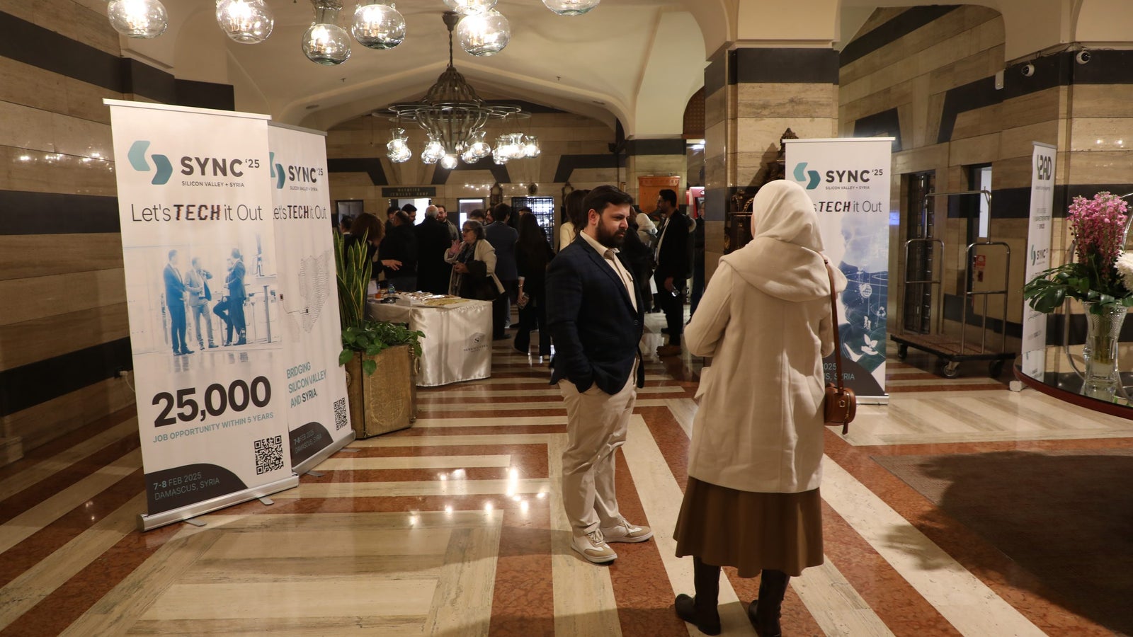 A spacious event venue with decorative lighting, featuring several banners promoting "SYNC 25" and "Let's TECH it Out." Attendees are engaged in conversation, and a woman in a light coat and headscarf is speaking to a man in a suit, while others mingle in the background.