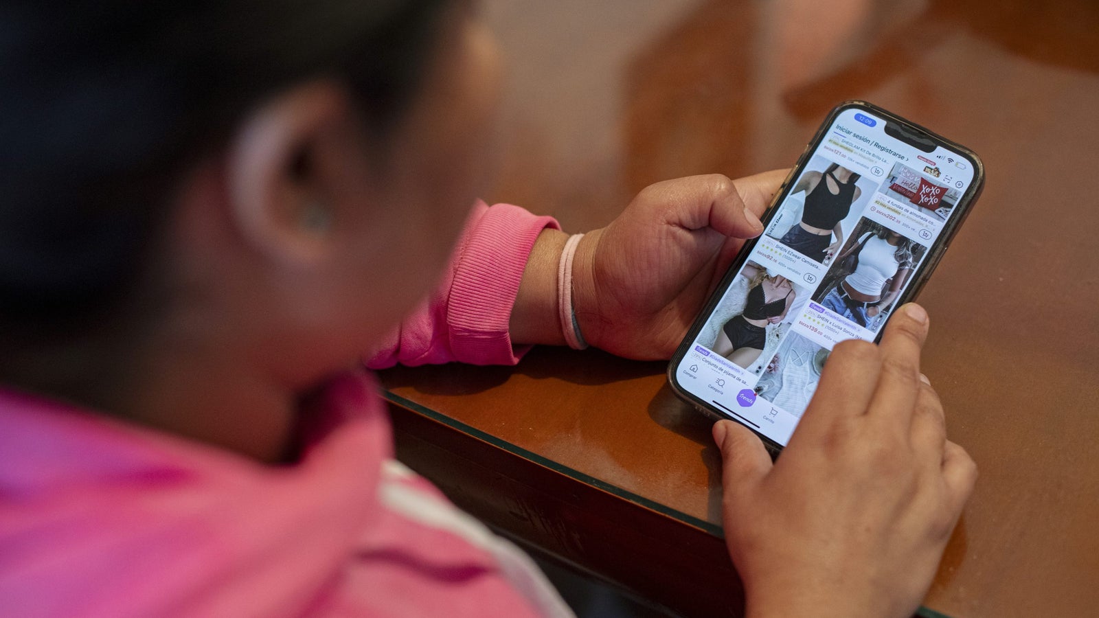 A person wearing a pink sweatshirt is holding a smartphone, browsing online shopping pages that display images of various tops or undershirts on a wooden table.