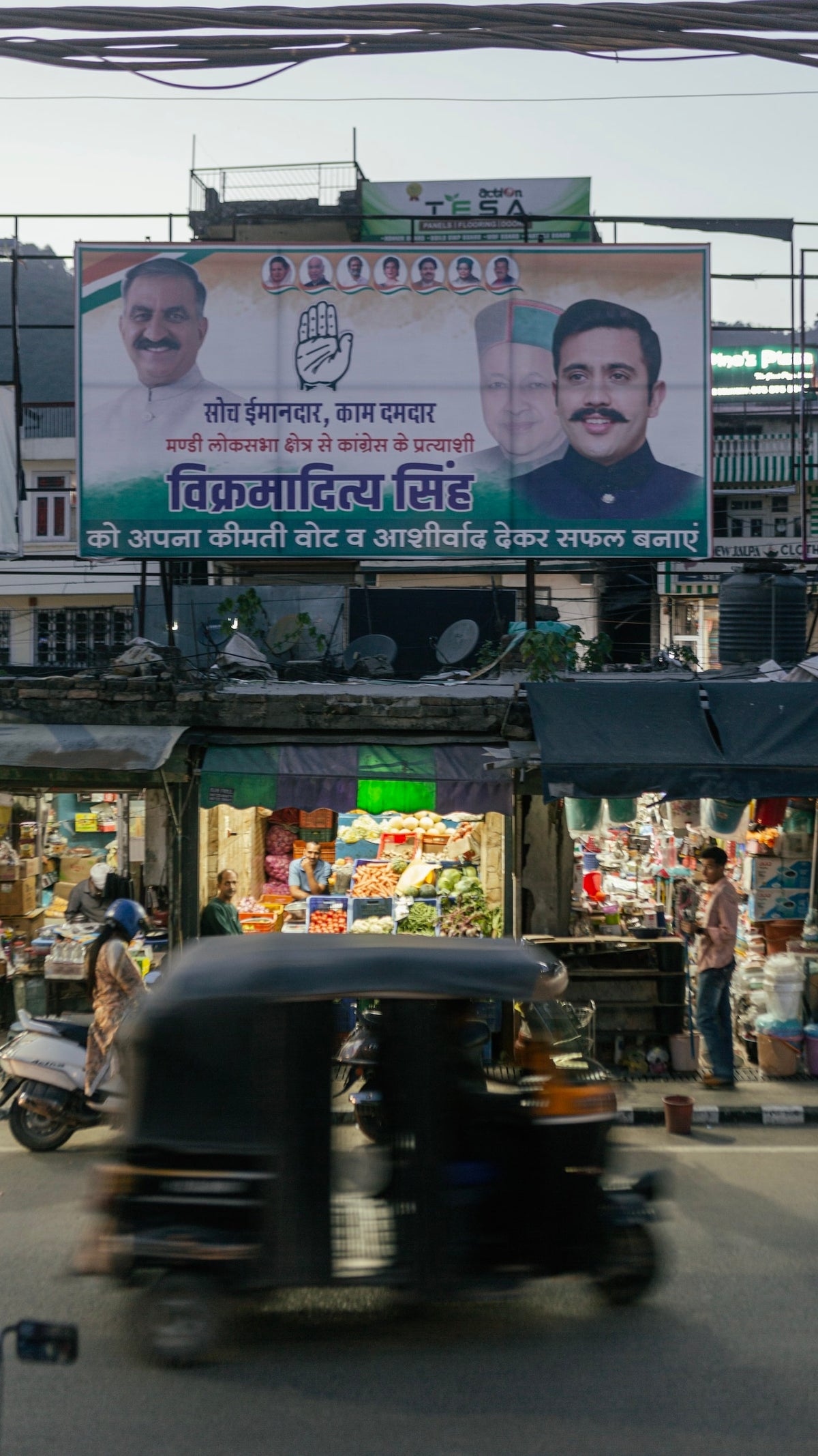 A photograph showing an Indian political poster hanging over a market in a city, as a rickshaw drives by.