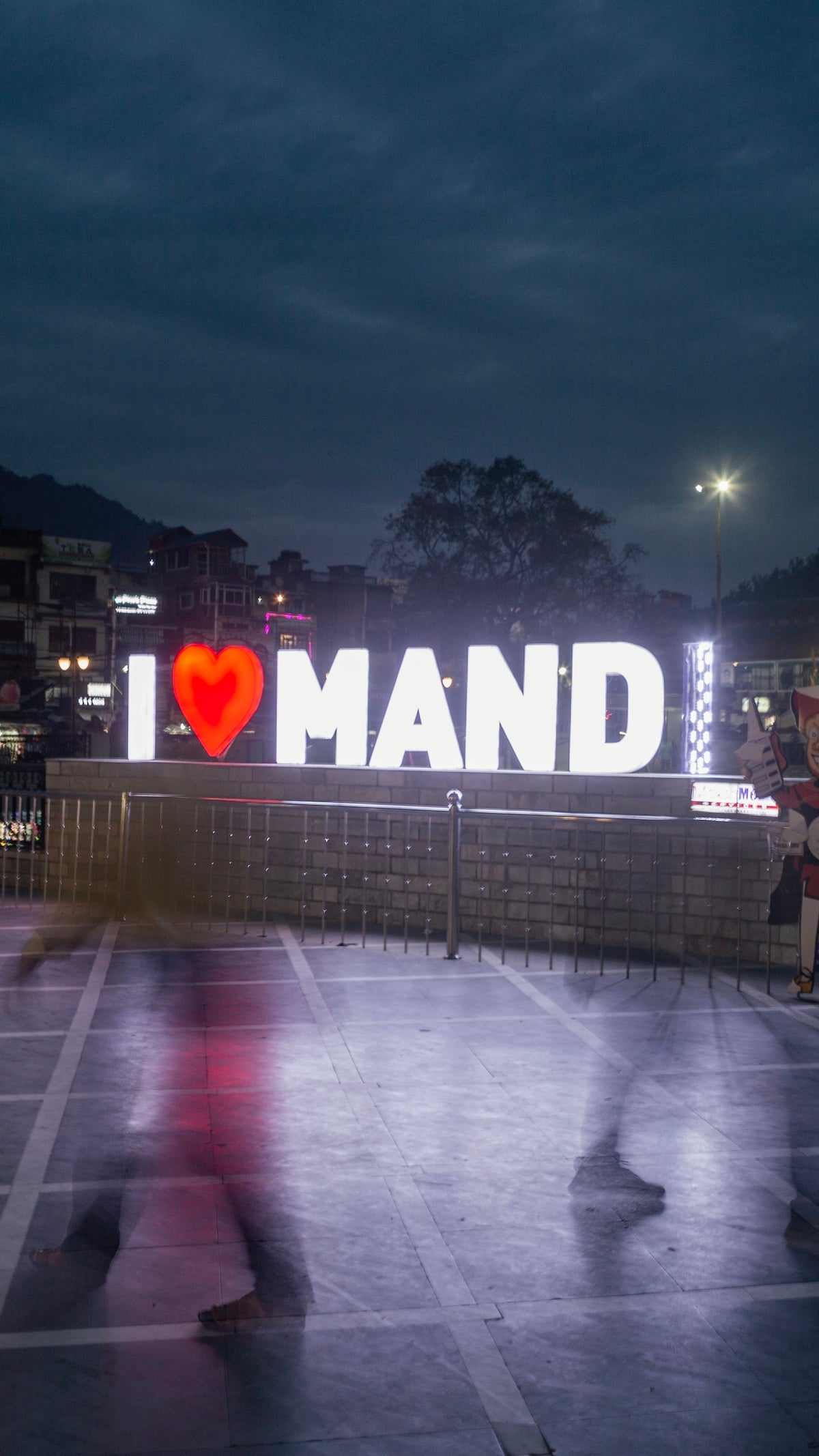 A photograph taken at night showing the blurred movement of people passed an "I love Mandi" sign in bright lights.
