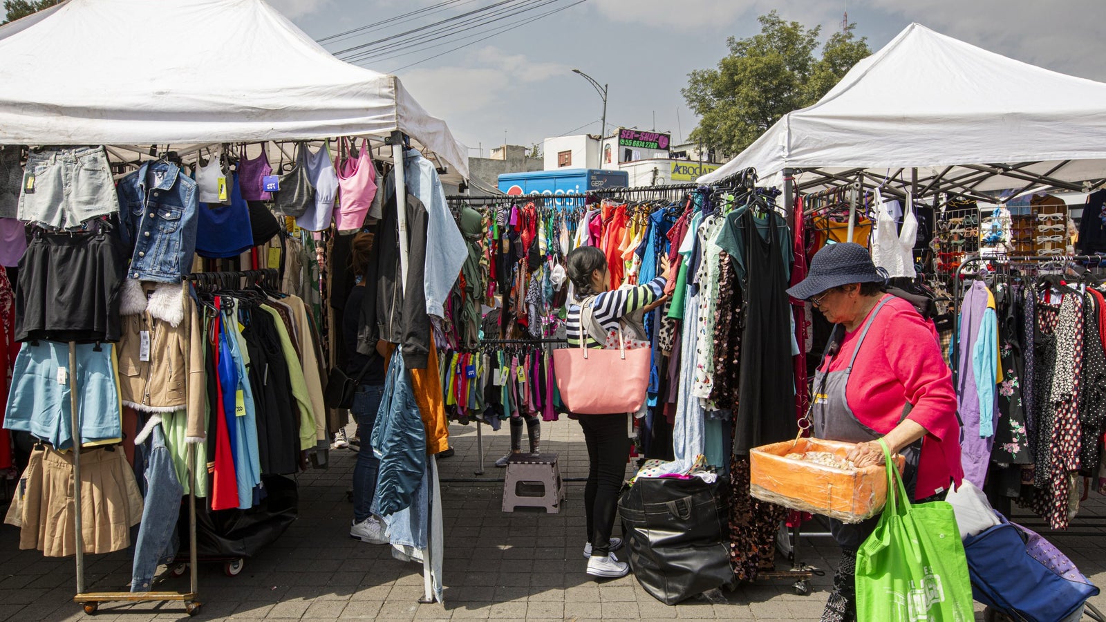 A busy outdoor market scene with two women shopping among colorful clothing racks; one woman with a pink bag admires a dress, while another older woman carries a basket and a green bag, surrounded by tents and a variety of clothes hanging on metal racks.