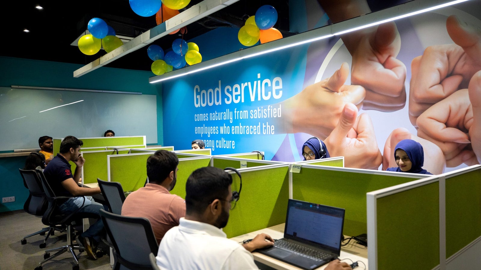 A photo showing a row of small desk spaces with people working in an office environment. Printed on the wall in the background is a picture of people's thumbs and text that reads "Good service comes naturally from satisfied employees who embraced the company culture."