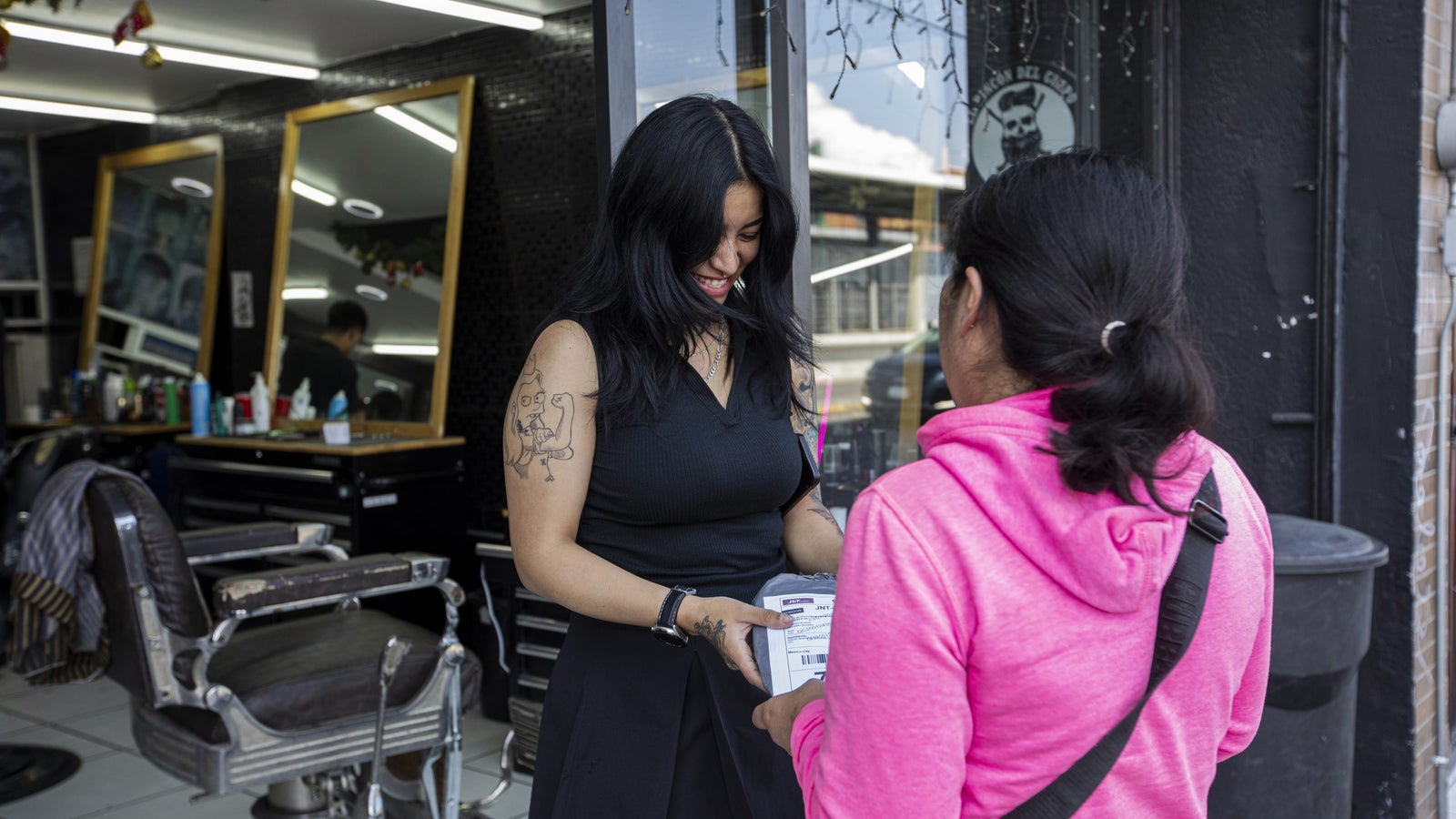 A woman with long black hair and a tattoo on her arm smiles while handing a package to another woman wearing a pink hoodie outside a barbershop, which features large mirrors and barber chairs in the background.