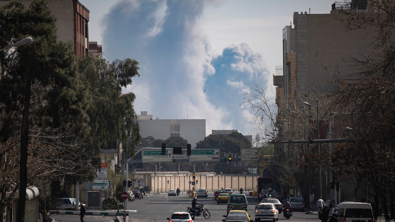 Smoke billows from buildings in the distance, viewed from a city street with vehicles and trees lining the road.