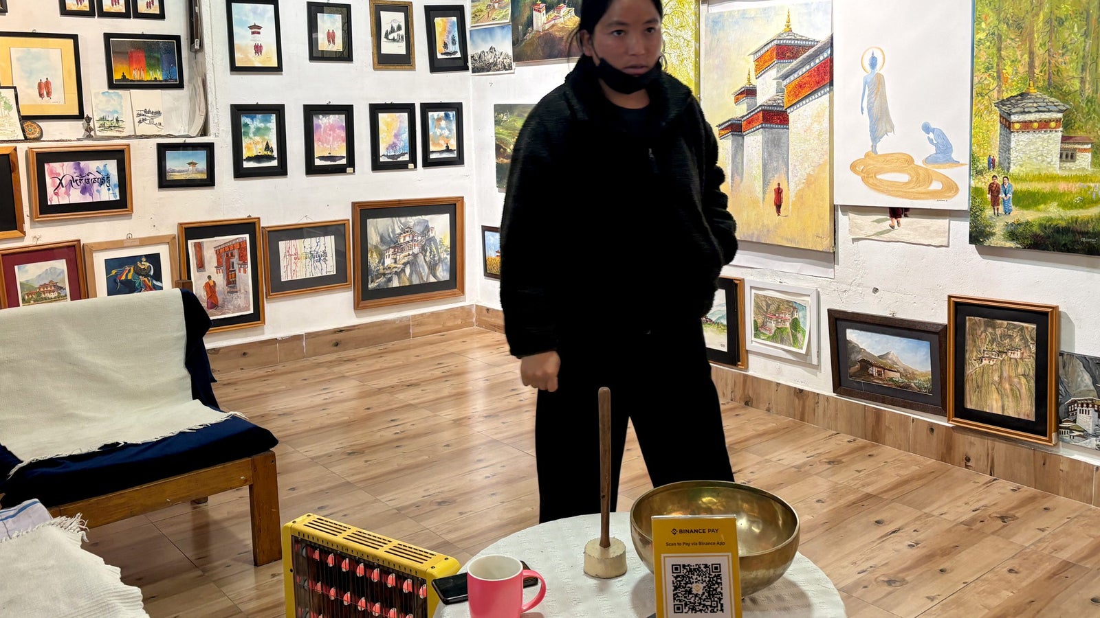 A person stands in an art gallery surrounded by various framed paintings, with a singing bowl and colored sticks on a table.