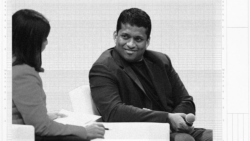 A man in a black blazer smiles while holding a microphone, engaging in conversation with a woman in a blue blazer sitting across from him.