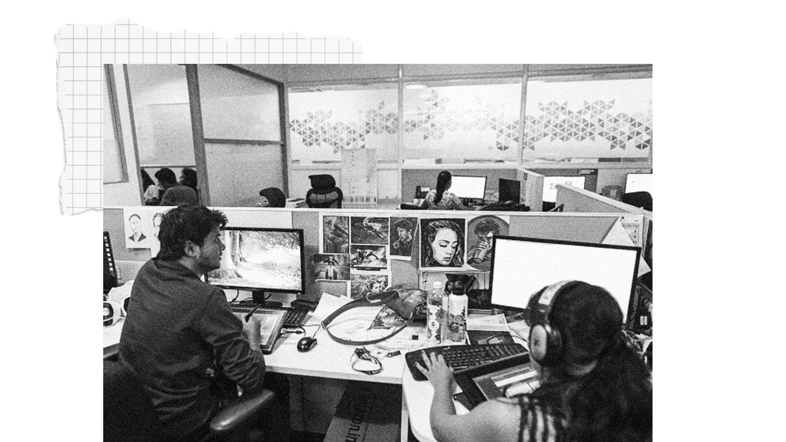 A workspace featuring two individuals at desks with dual monitors, focused on their screens. The man on the left is using a drawing tablet, while the woman on the right wears headphones and uses a keyboard. The desks are decorated with various artworks and personal items. Several other people are visible in the background working at additional desks.