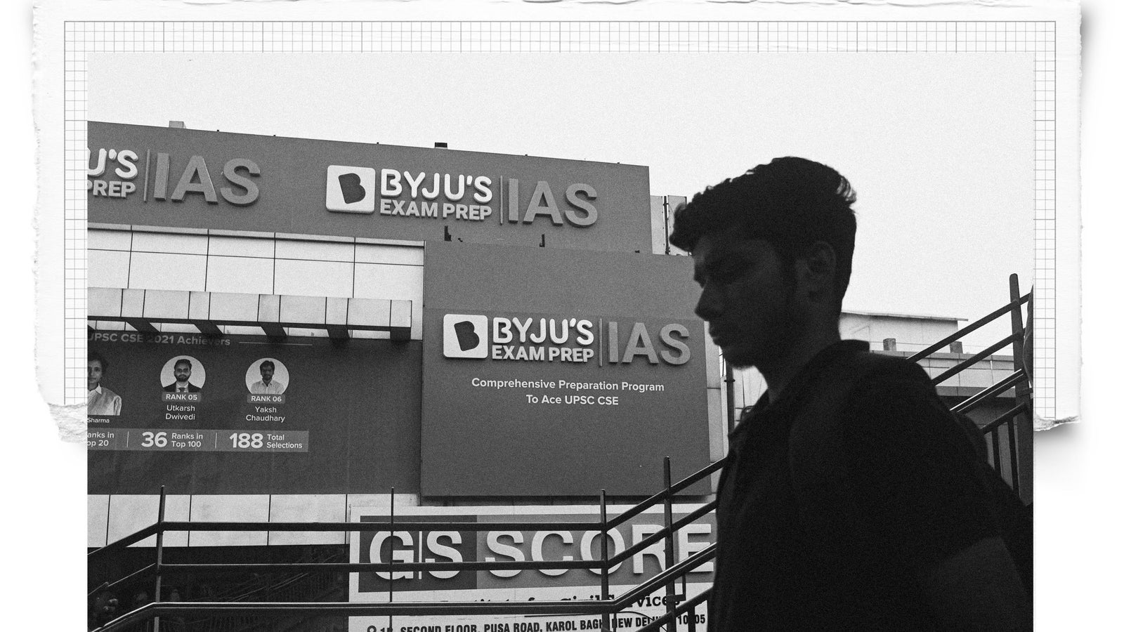 A silhouette of a young man walking past a large advertisement for BYJU'S Exam Prep, which emphasizes its IAS preparation program with text outlining program details and achievements of students, set in a black and white tone.